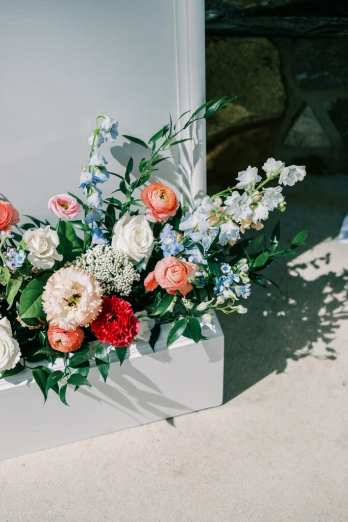 Close-up of floral arrangement at wedding welcome sign – A stunning floral arrangement featuring white roses, coral ranunculus, and blue delphiniums at the base of the wedding welcome sign at The Historic Robertson House.