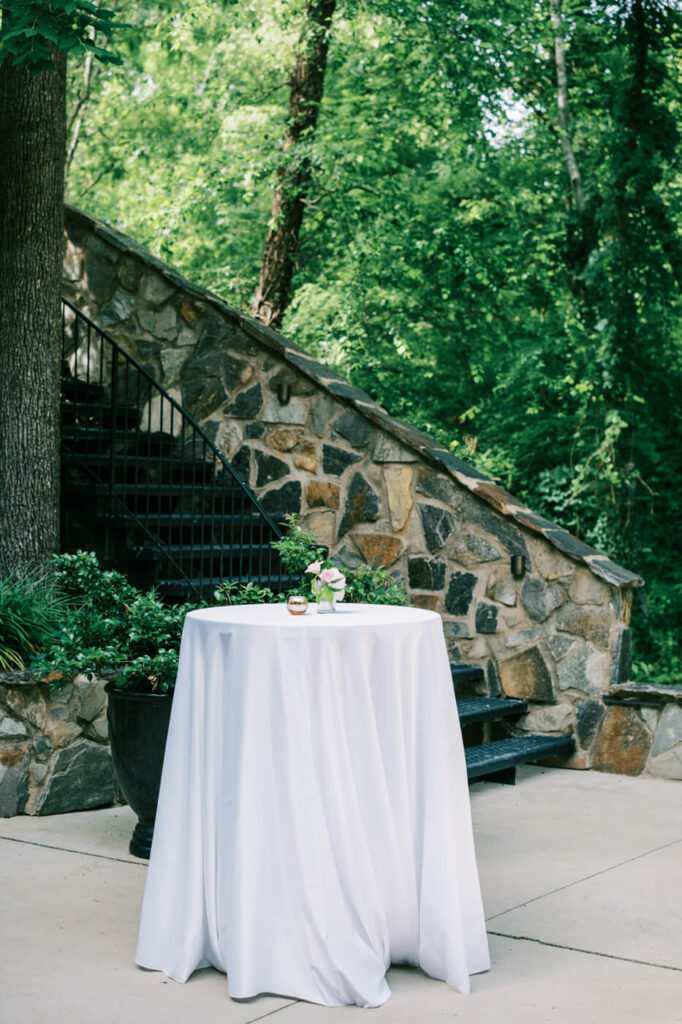 Intimate cocktail hour setup at a garden wedding – A round cocktail table draped in white linen, adorned with delicate floral accents, placed near a rustic stone staircase in the lush gardens of The Historic Robertson House.