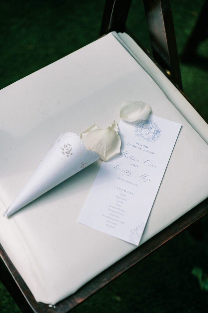 Wedding program and rose petal cone on ceremony chair – A personalized wedding program with an elegant monogram, placed on a wooden ceremony chair with white petals in a lace-trimmed cone at a garden wedding.