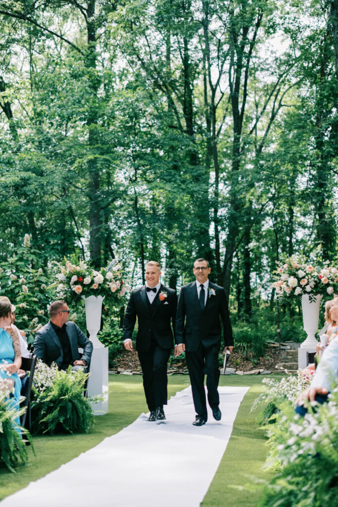 Groom walking down the aisle with officiant at outdoor wedding – The groom, dressed in a classic black tuxedo with a coral boutonniere, walks down the lush green aisle alongside the officiant at The Historic Robertson House.