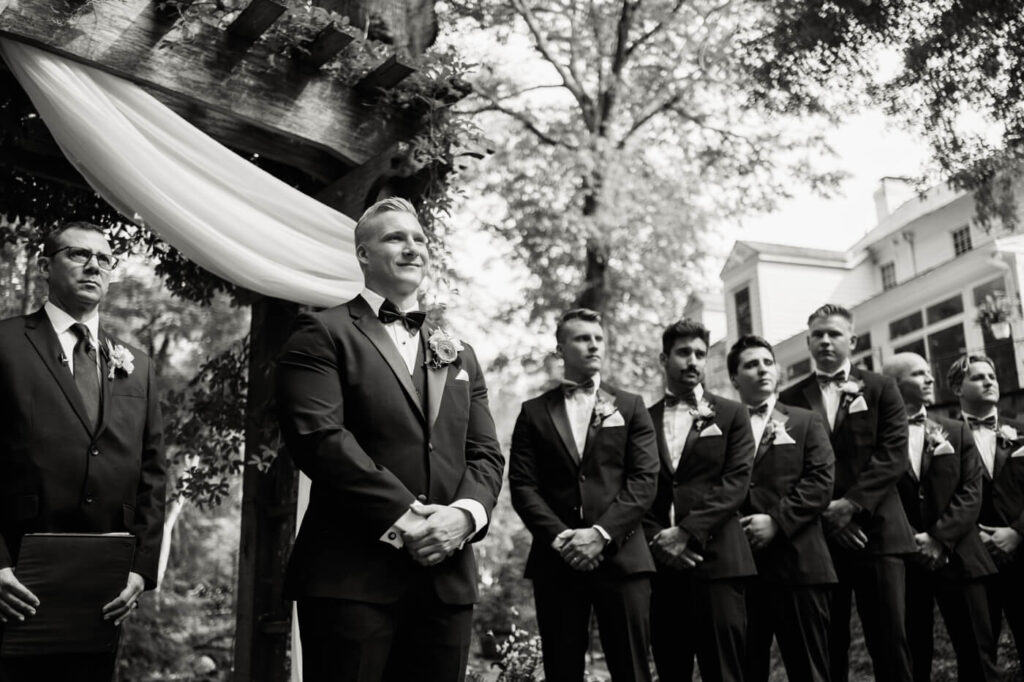 Groom awaiting the bride at a garden wedding altar – A black-and-white image capturing the groom’s anticipation as he stands beneath a wooden arbor draped with white fabric, surrounded by his groomsmen.