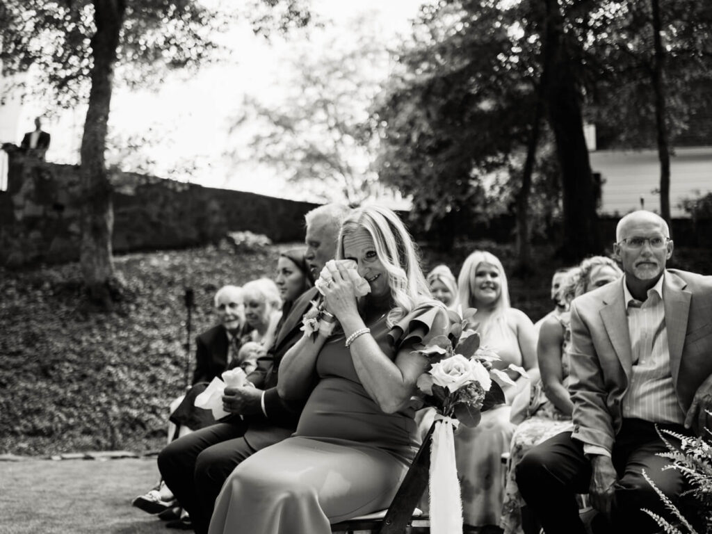 Mother of the groom wiping away tears during wedding ceremony – A touching black-and-white moment of the groom’s mother wiping away tears while seated among wedding guests at an emotional garden ceremony.