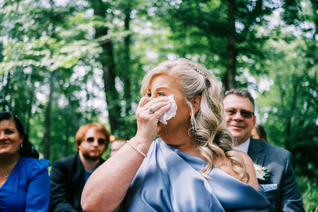 Mother of the bride overcome with emotion during ceremony – mother in a dusty blue gown dabs her eyes with a tissue as she watches the couple exchange vows in a picturesque outdoor setting.