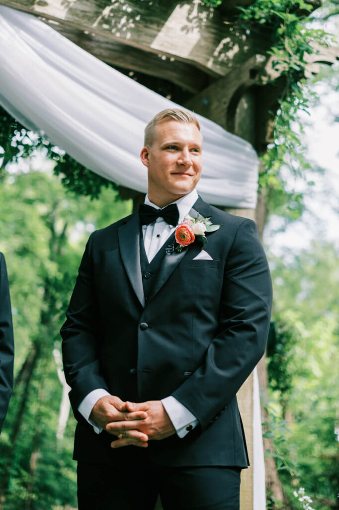 Groom's reaction to seeing the bride at the altar – A heartfelt moment as the groom, dressed in a black tuxedo with a classic black bowtie, smiles in anticipation while standing under a greenery-wrapped wooden pergola.