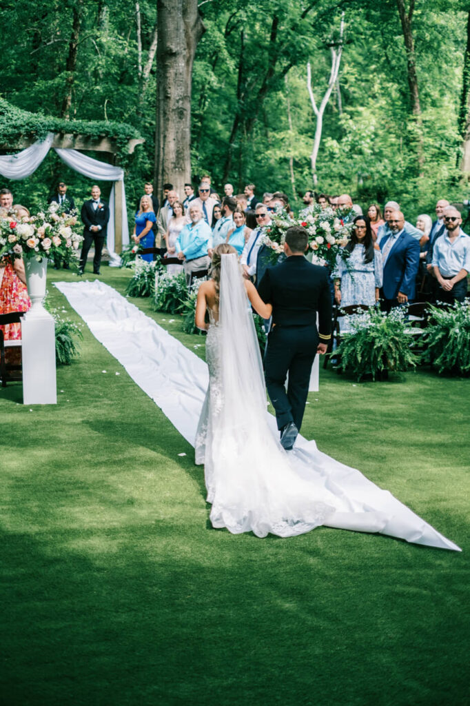 Bride walking down the aisle with her brother at The Historic Robertson House wedding – The bride, in a stunning lace wedding gown, walks down the aisle with her brother in a military uniform, surrounded by floral arrangements and lush greenery.