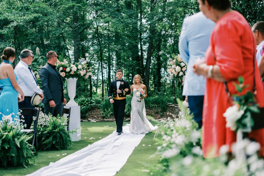 Bride and brother making their way down the aisle at an elegant outdoor wedding – The bride, holding a bouquet of blush and blue flowers, walks arm-in-arm with her brother in a military dress uniform during a romantic outdoor ceremony.