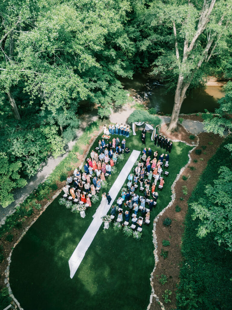 Aerial view of an outdoor wedding ceremony at The Historic Robertson House – A breathtaking drone shot captures the entire wedding ceremony site, featuring a floral-lined aisle, seated guests, and a scenic creek running beside the venue.