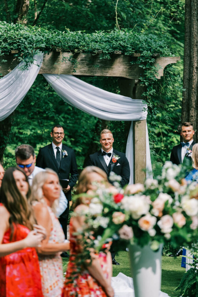 Groom's first look at the bride walking down the aisle at The Historic Robertson House – The groom, in a classic black tuxedo, beams with joy as he sees his bride walking toward him under the ceremony arbor draped with white fabric.