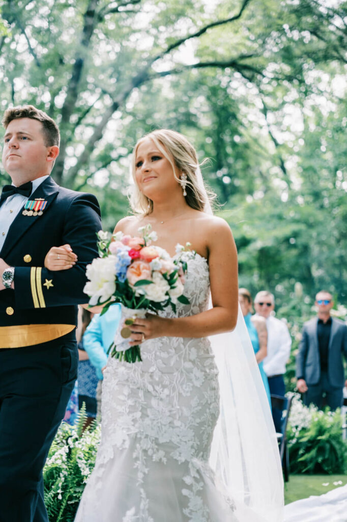 Bride emotional and brother making their way down the aisle at an elegant outdoor wedding – The bride, holding a bouquet of blush and blue flowers, walks arm-in-arm with her brother in a military dress uniform during a romantic outdoor ceremony.