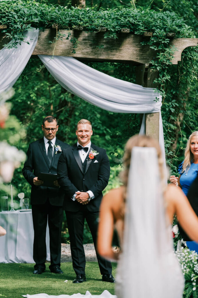 Groom smiling as his bride walks down the aisle at The Historic Robertson House – The groom stands beneath a greenery-covered wooden arbor, waiting for his bride with a heartfelt expression.