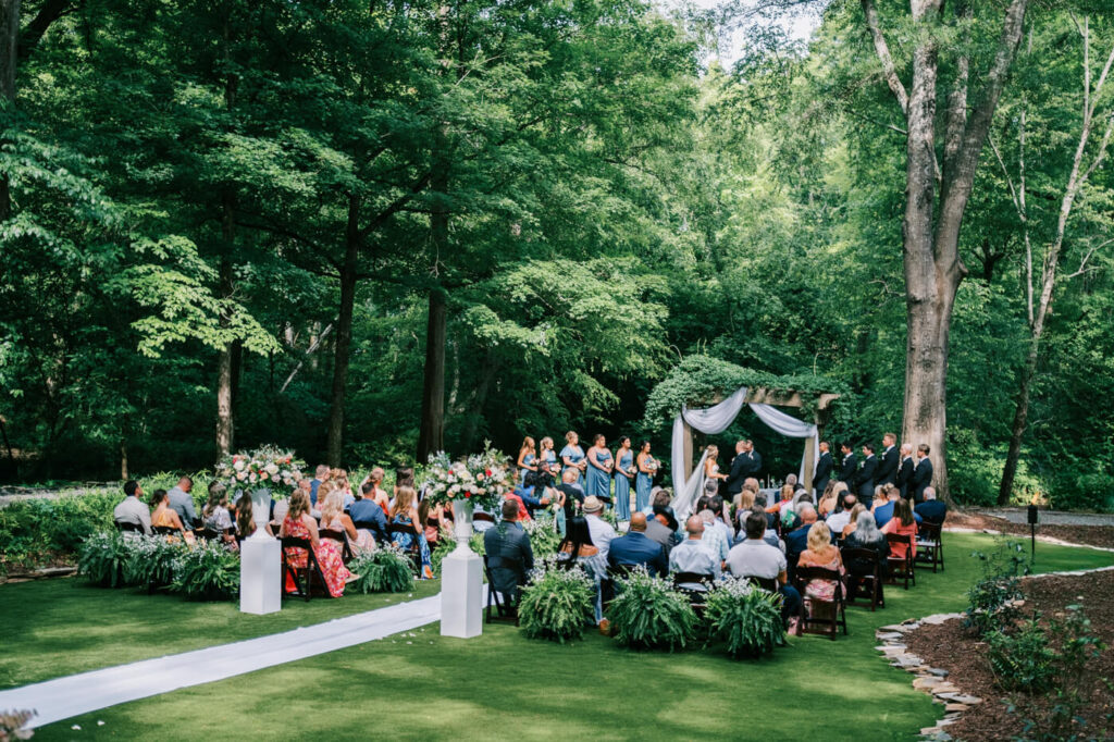 Outdoor wedding ceremony surrounded by greenery at The Historic Robertson House – Guests sit beneath towering trees, witnessing the couple exchange vows in a natural and romantic woodland setting.