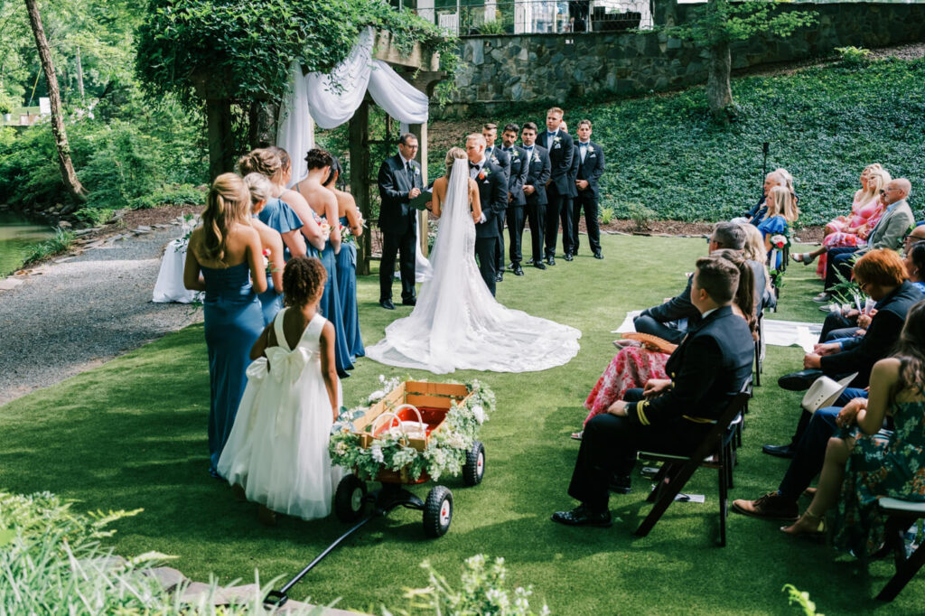 Ring bearer in a decorated wagon during a wedding ceremony at The Historic Robertson House – A young flower girl in a white dress stands next to a floral-adorned red wagon, adding a sweet and whimsical touch to the ceremony.