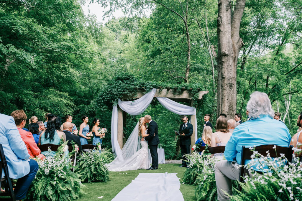 Bride and groom share a romantic first kiss at their Historic Robertson House wedding ceremony, surrounded by lush greenery and elegant white drapery.