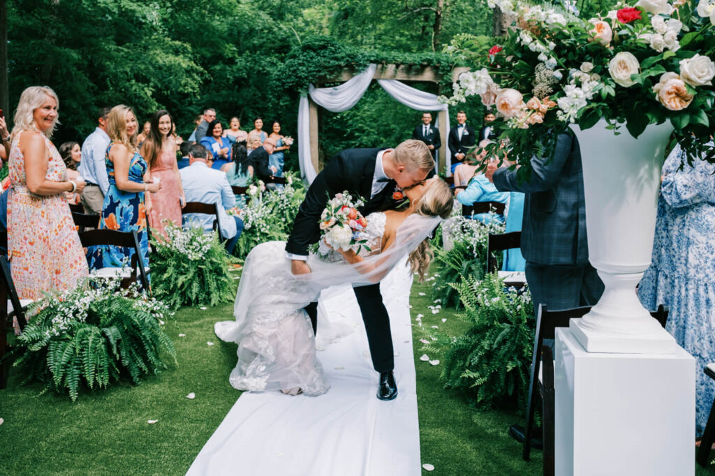 Bride and groom share a dramatic dip kiss surrounded by lush floral arrangements and happy guests at their garden wedding ceremony.