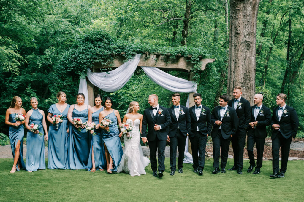 Bridal party poses in front of the ceremony arbor, bridesmaids in elegant blue satin dresses and groomsmen in classic black tuxedos.