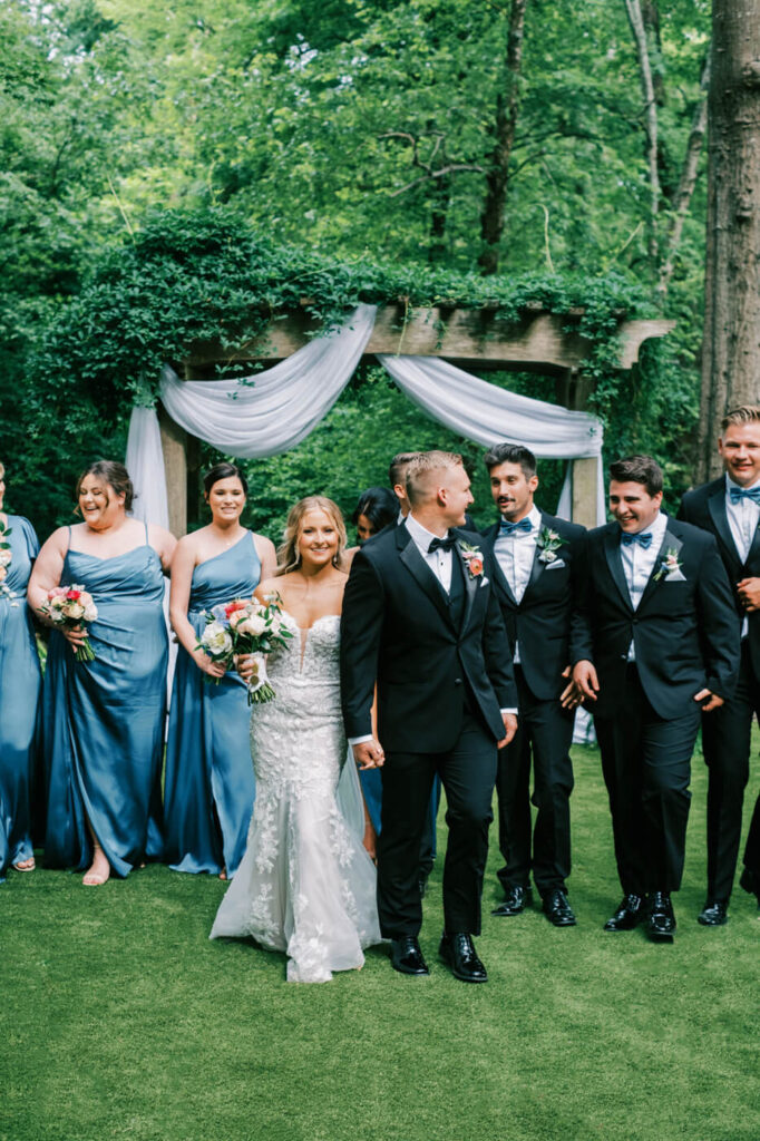 Candid moment of the bride and groom walking hand in hand with their bridal party, laughing and celebrating in the garden setting.