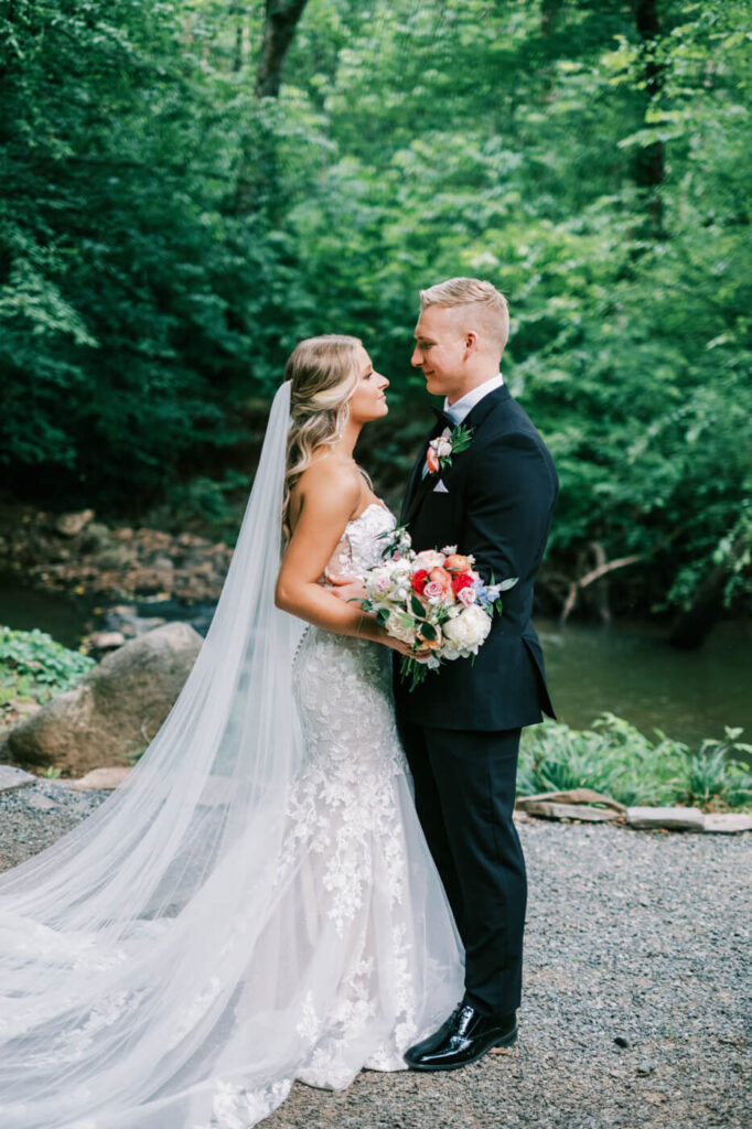 Bride and groom stand together by the riverside, gazing into each other's eyes, the bride holding a vibrant bouquet of pink and white roses.