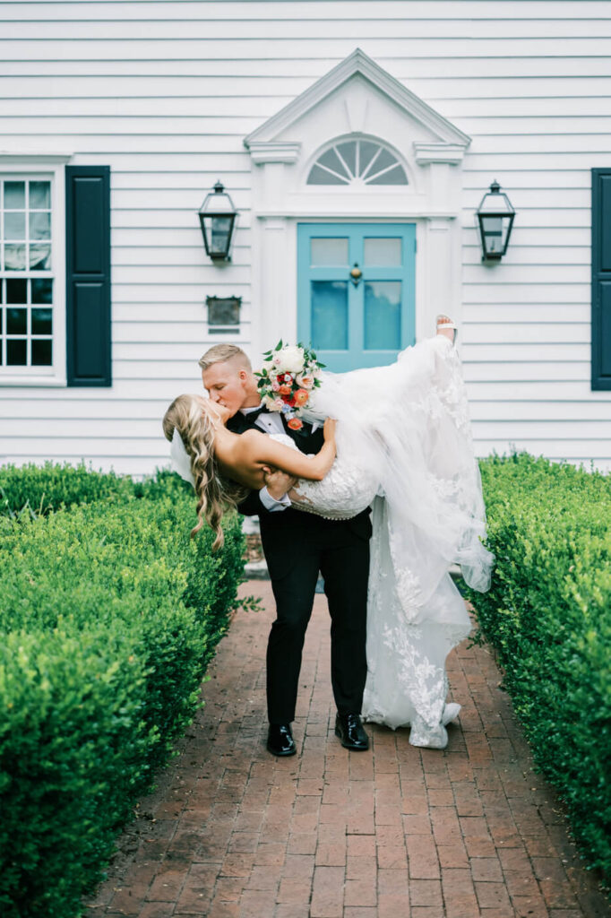 Groom carrying bride in front of The Historic Robertson House – A romantic wedding portrait of a groom lifting his bride in front of a classic white colonial home with blue doors at The Historic Robertson House.