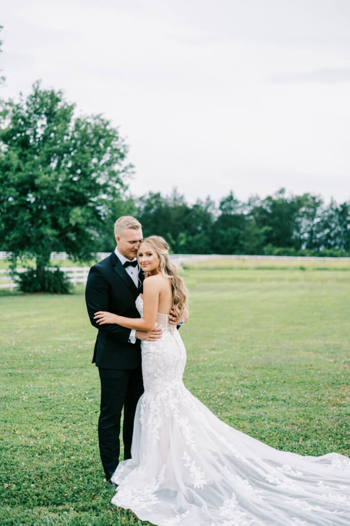 Bride and groom embrace in an open field – A newlywed couple shares an intimate moment in a lush green field, with the bride’s lace wedding gown elegantly trailing behind her.