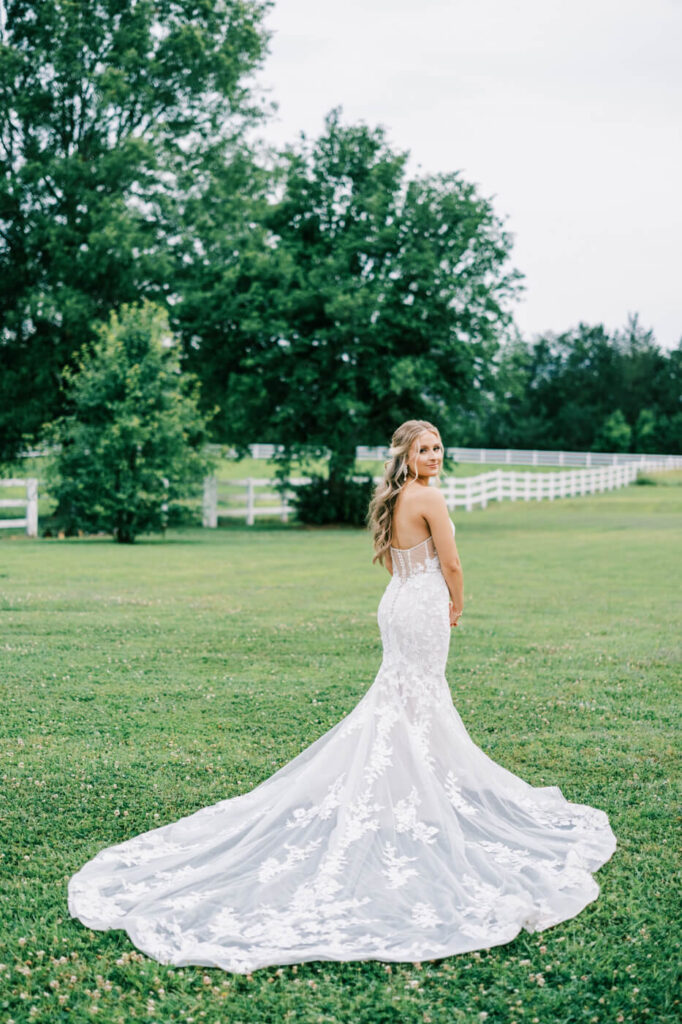 Bride standing alone in a scenic meadow – A stunning bridal portrait featuring the bride in a fitted lace gown, gazing back over her shoulder with rolling pastures in the background.