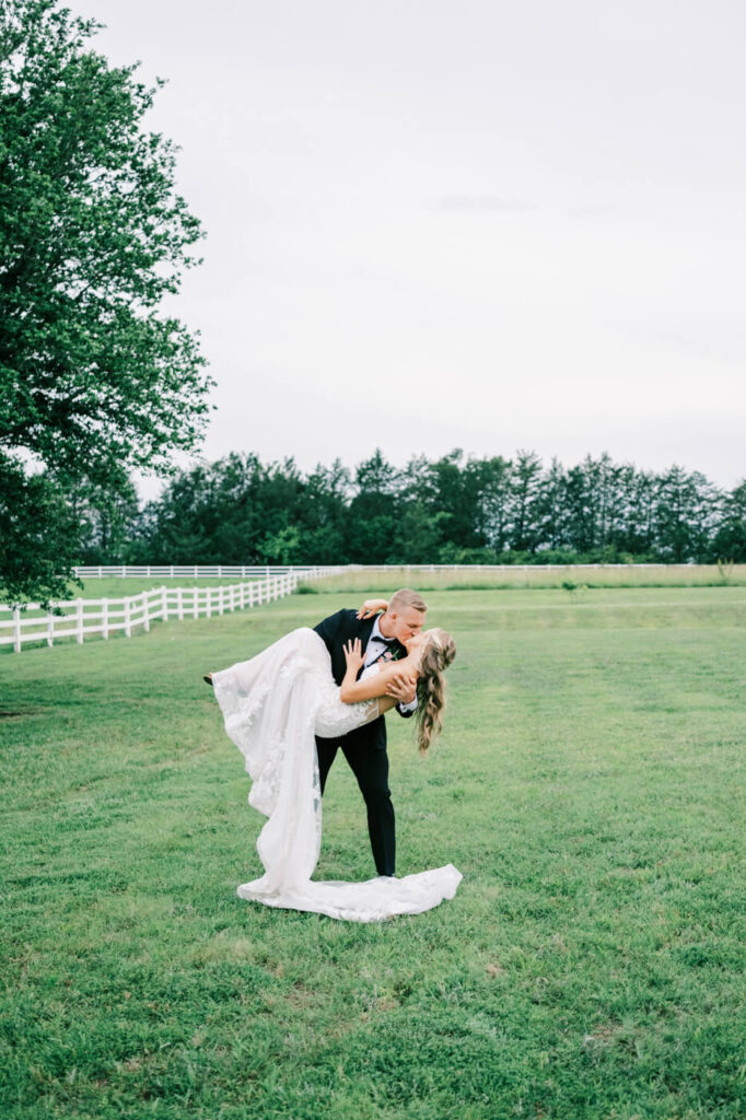 Groom dips bride for a romantic kiss in the countryside – A cinematic wedding photo of the groom sweeping his bride off her feet, kissing her in the middle of a green pasture with a white fence in the background.