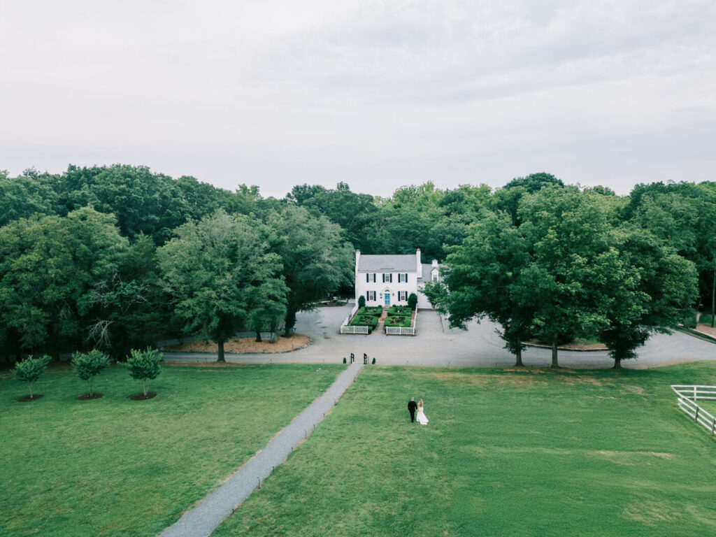 Aerial view of The Historic Robertson House and newlywed couple – A breathtaking drone shot capturing the expansive grounds of The Historic Robertson House with the bride and groom walking hand in hand along the pathway.