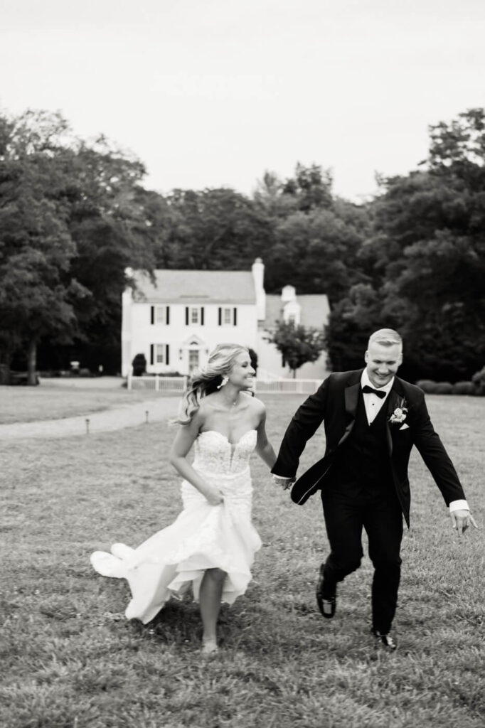 Black and white wedding portrait of couple running – A joyful moment as the newlyweds run across the grass, laughing, with the elegant Historic Robertson House in the background.