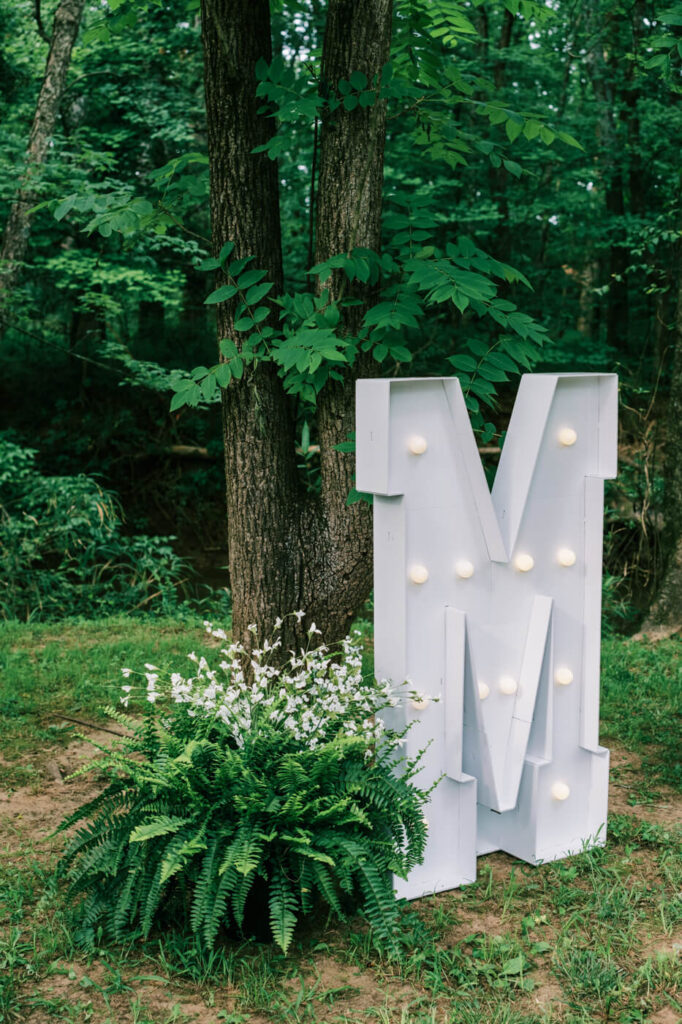 Illuminated wedding marquee letter ‘M’ in the woods – A personalized wedding detail featuring a large light-up ‘M’ marquee nestled in the greenery, surrounded by ferns and wildflowers.