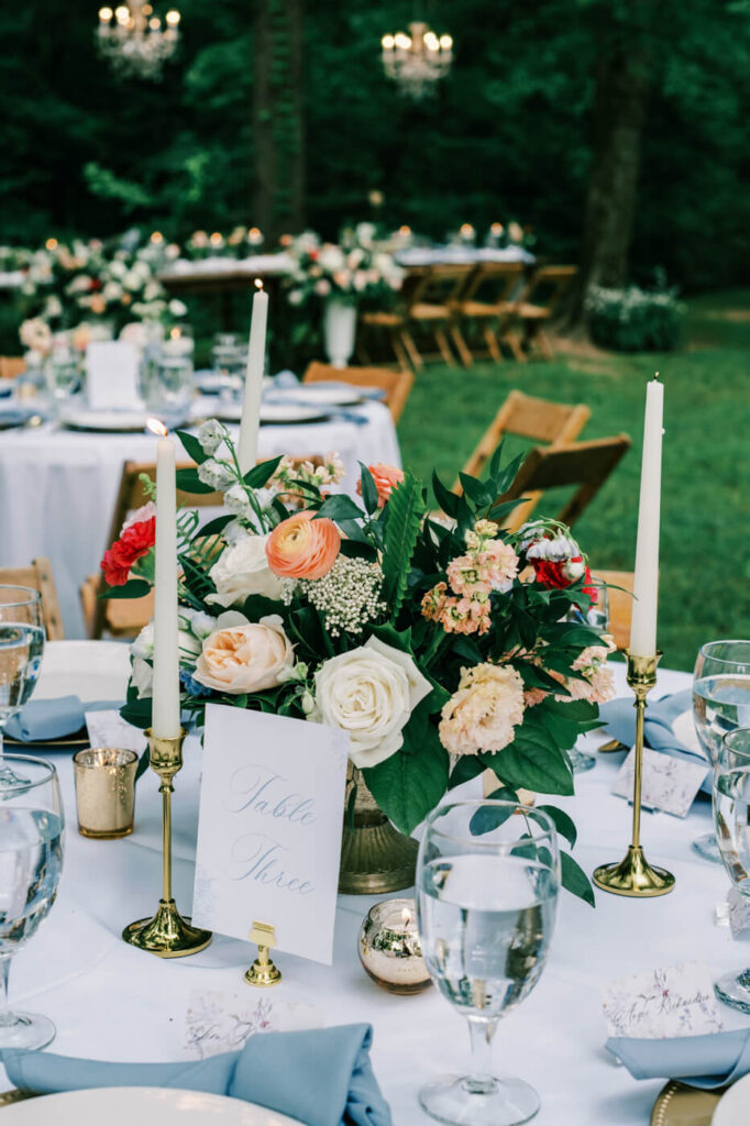 Elegant wedding reception table at The Historic Robertson House – A beautifully set round table with gold-rimmed plates, soft blue napkins, tall taper candles, and a lush floral centerpiece of peach, white, and red roses, set against the enchanting wooded backdrop.