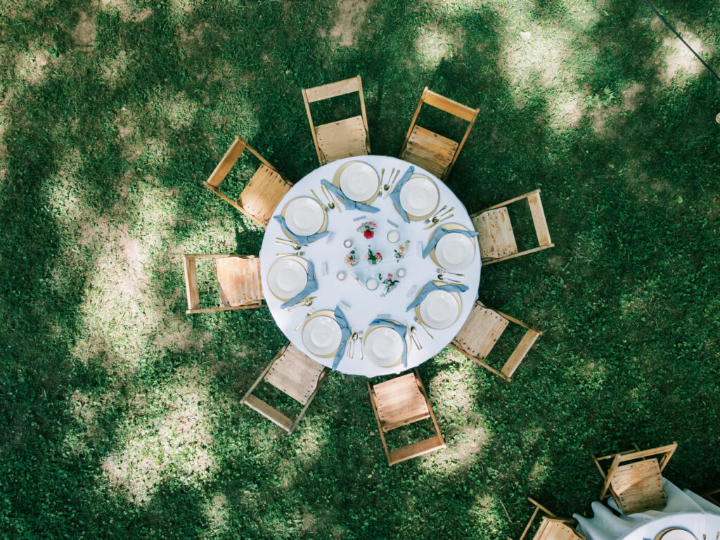 Overhead view of an intimate wedding table setup – A circular wedding reception table with wooden chairs, fine china, gold flatware, and delicate floral arrangements, surrounded by dappled sunlight filtering through the trees.