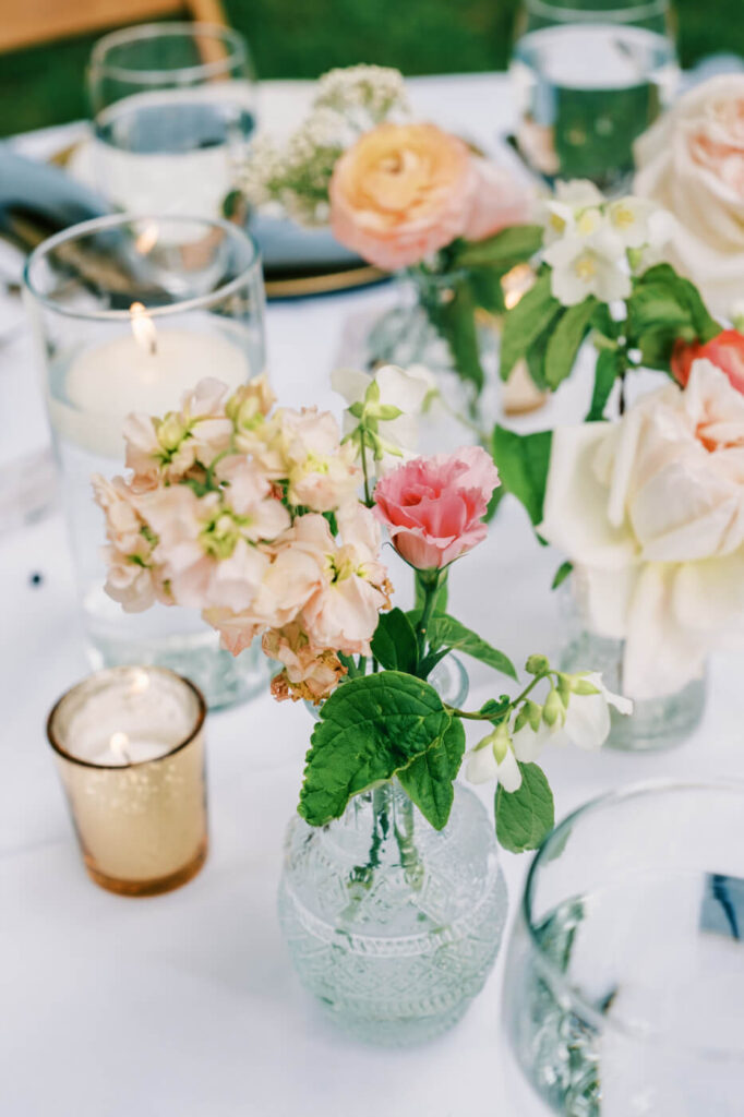 Romantic floral arrangement at a garden wedding reception – A close-up of a delicate pink and white flower arrangement in a vintage glass vase, accompanied by glowing candles on a softly lit reception table.