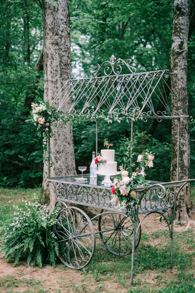 Vintage wedding cake cart with floral décor – A charming antique-inspired metal cart adorned with greenery and florals, showcasing a classic white wedding cake with elegant gold accents, nestled in a serene forest reception area.