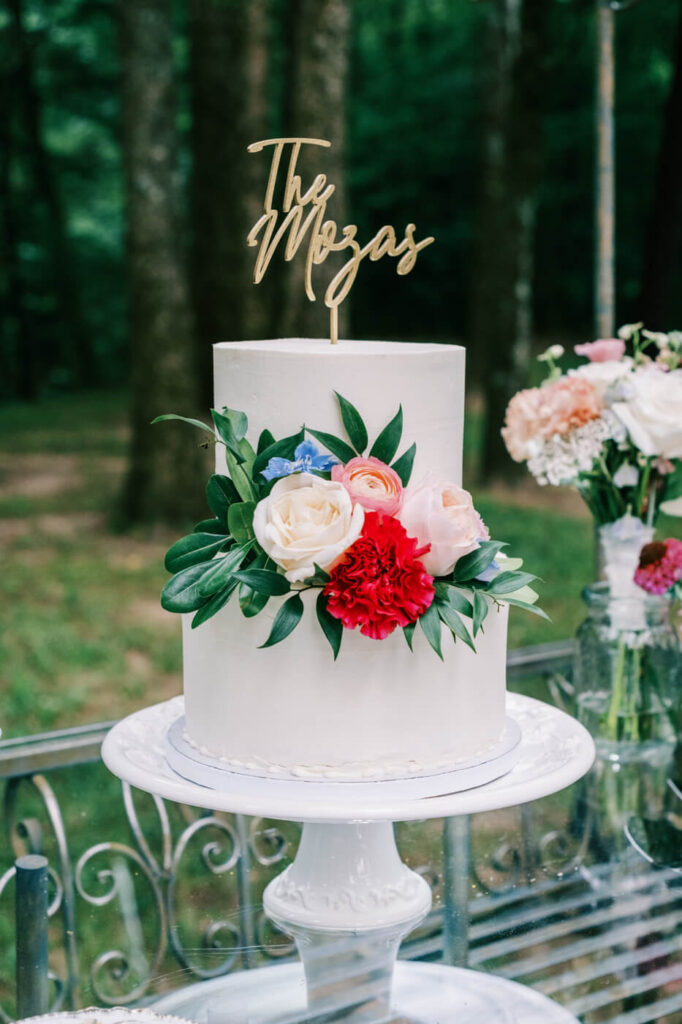 Minimalist white wedding cake with floral accents – A simple yet sophisticated single-tier white cake adorned with fresh roses, greenery, and a custom gold topper, perfectly complementing the rustic garden wedding setting.