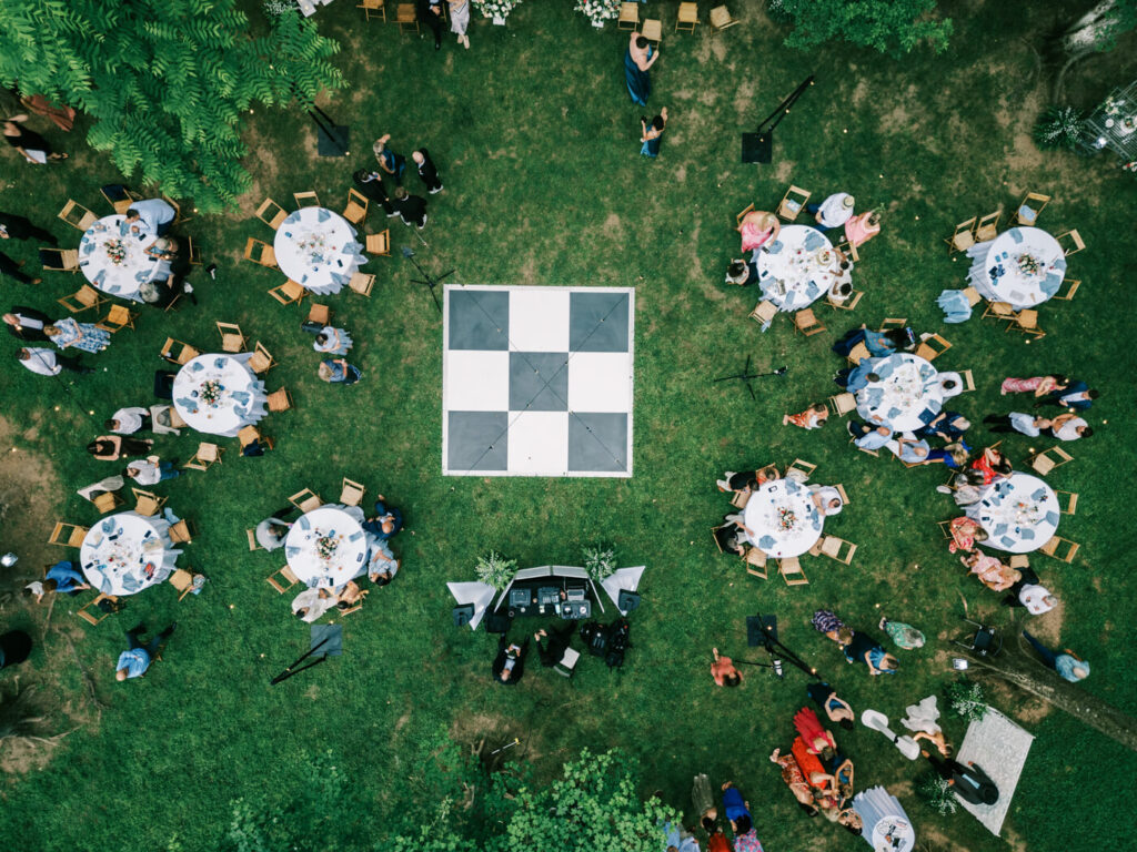 Aerial view of an enchanting forest wedding reception – A stunning drone shot capturing the wedding reception layout, featuring round tables, wooden chairs, a black-and-white dance floor, and guests celebrating under string lights.