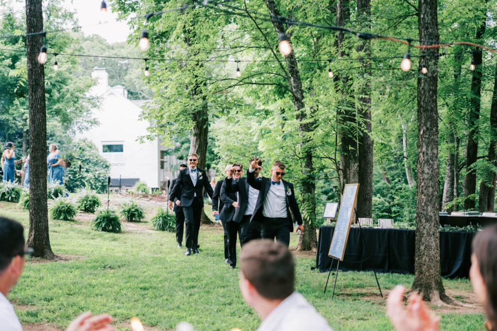 Groomsmen making a grand entrance at a forest wedding – A stylish group of groomsmen dressed in classic black tuxedos and bow ties, walking confidently towards the reception area, surrounded by lush greenery and twinkling string lights.