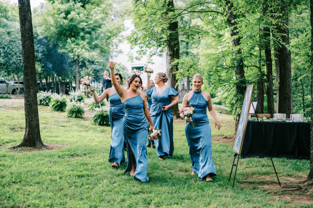 Bridesmaids' Grand Entrance at The Historic Robertson House – A group of bridesmaids in elegant blue gowns joyfully enter the wedding reception, holding bouquets, with lush greenery surrounding them at The Historic Robertson House.