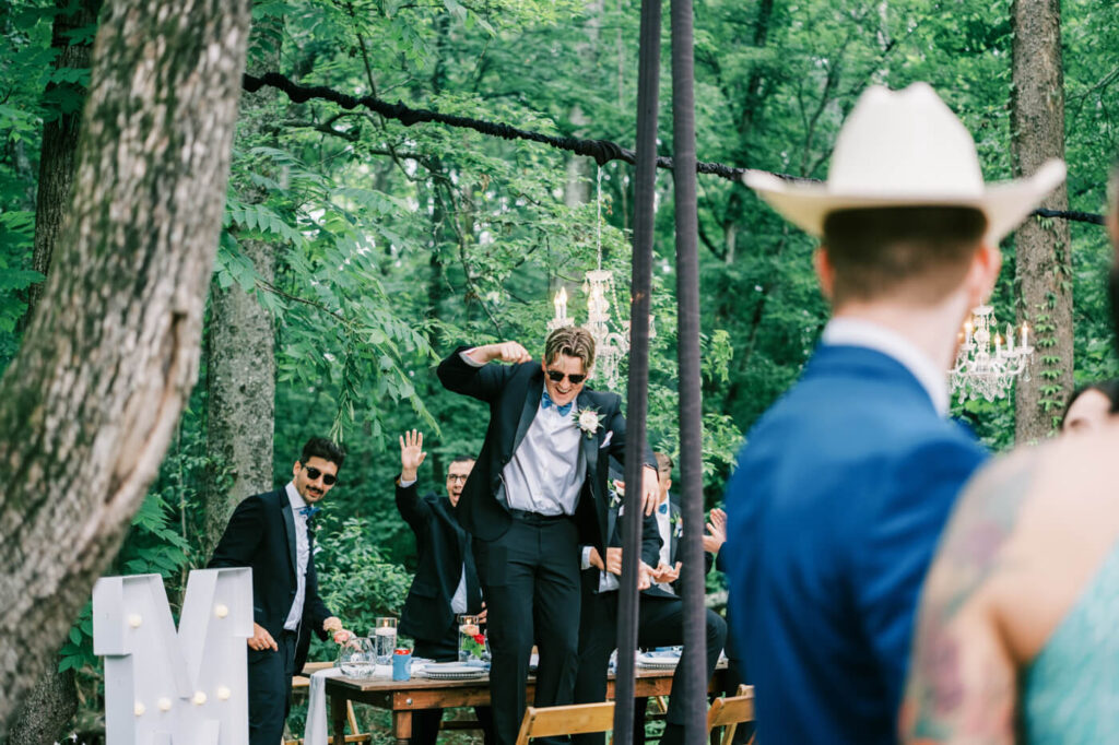 Groomsmen Celebrating at Outdoor Wedding Reception – A groomsman in a black tuxedo with a blue bow tie stands on a chair dancing, while his friends cheer him on at a forested outdoor wedding venue.