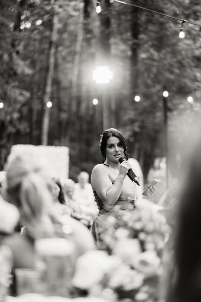 Bridesmaid Giving Heartfelt Speech at Wedding Reception – A bridesmaid in a satin blue gown delivers an emotional toast to the couple under the romantic glow of string lights at an intimate woodland wedding.