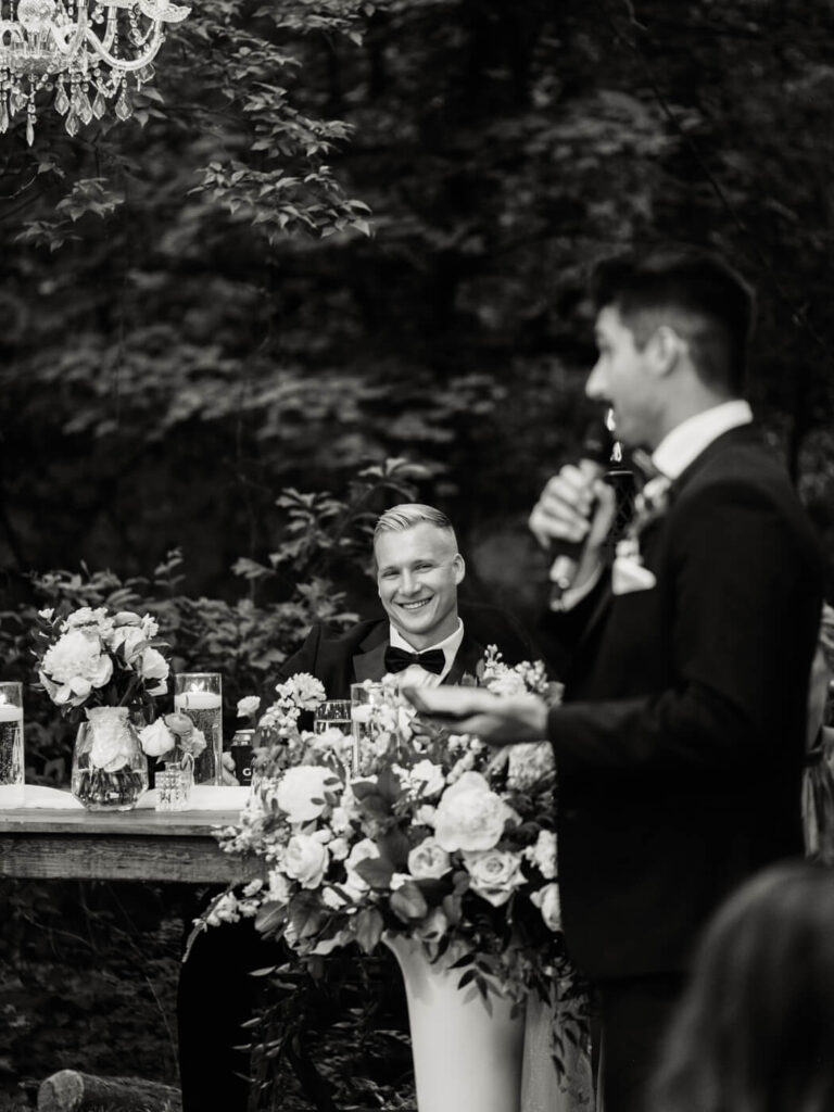 Groom Smiling During Best Man's Speech – A black and white candid moment of the groom grinning as his best man gives a heartfelt toast at their outdoor wedding reception.