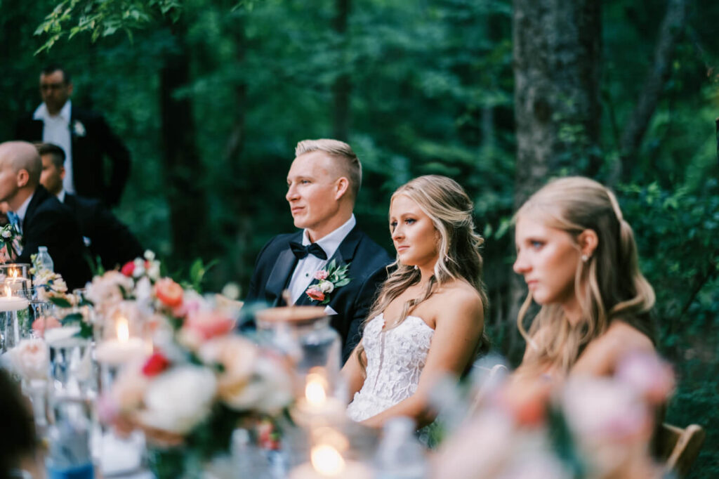 Bride and Groom Listening to Wedding Speeches – The bride and groom sit at their floral-adorned head table, gazing at their loved ones as they enjoy heartfelt speeches under a canopy of trees.