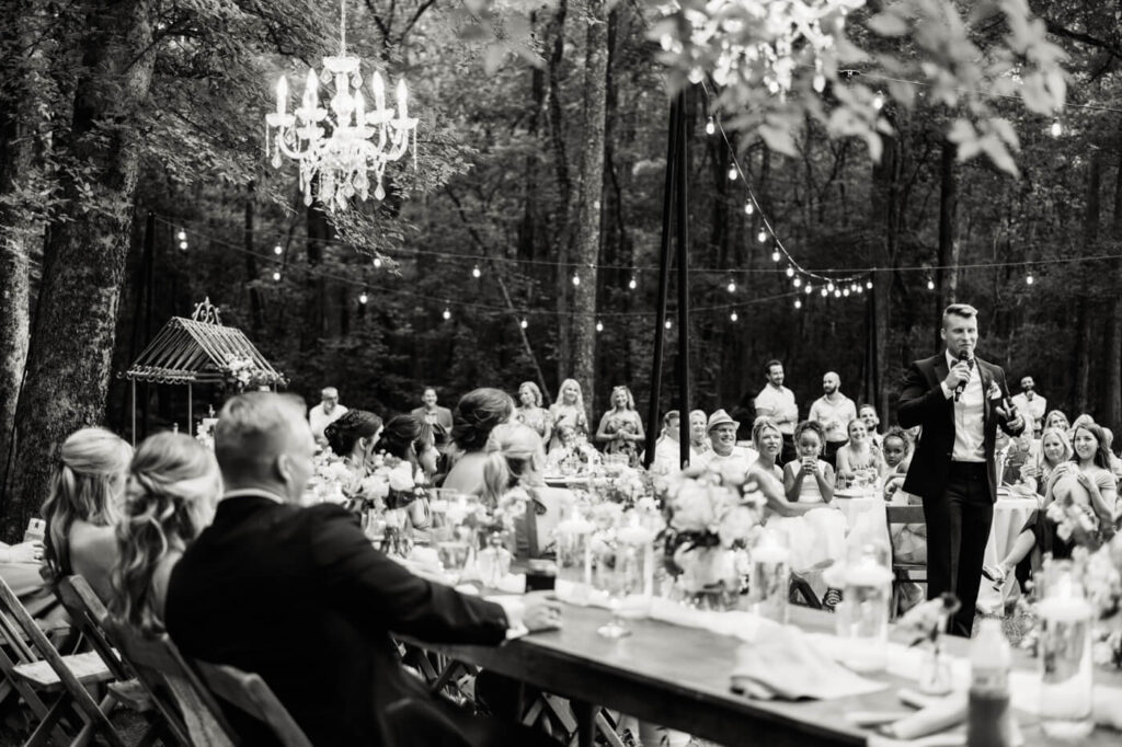 Romantic Outdoor Wedding Reception with Chandeliers and String Lights – A beautifully lit wedding reception featuring a groom giving a speech, with guests seated at long wooden tables surrounded by elegant florals.