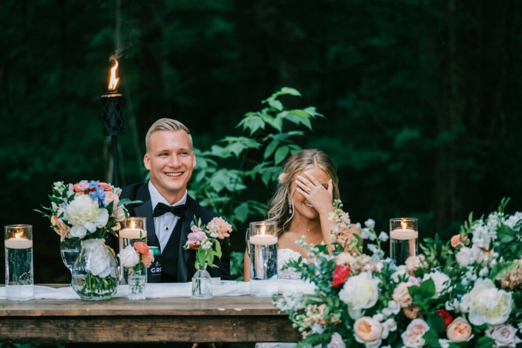 Bride Laughing at Wedding Toast with Groom Smiling – A joyful moment as the bride covers her face with laughter while her groom beams, seated at a candlelit sweetheart table adorned with pastel flowers.