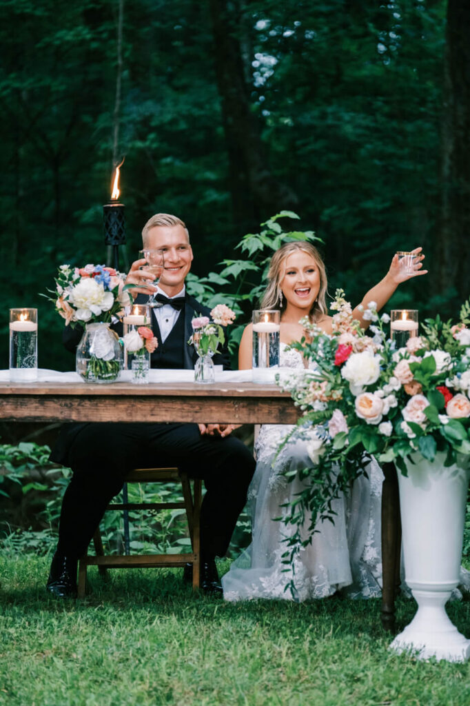 Bride and Groom Raising Glasses for Wedding Toast – The newlyweds celebrate with a toast, surrounded by lush floral arrangements, golden candlelight, and a romantic forest setting at The Historic Robertson House.