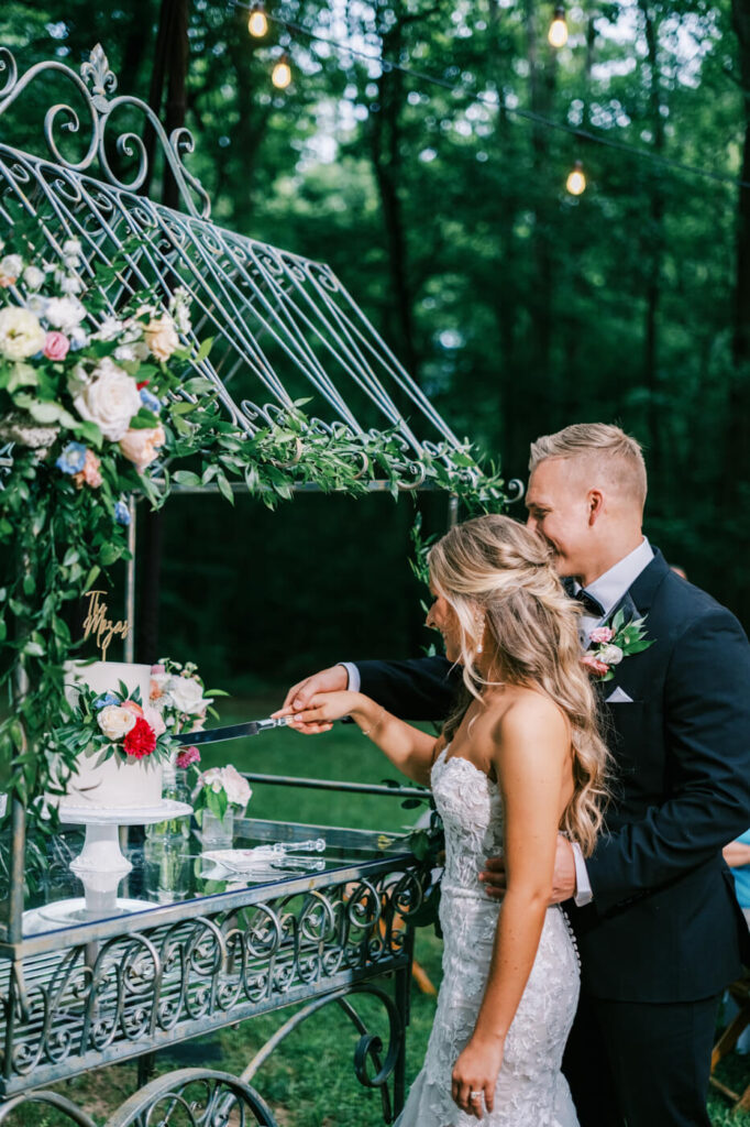 Bride and Groom Cutting Wedding Cake in Romantic Outdoor Setting – The couple shares a sweet moment cutting their elegant white wedding cake, adorned with fresh florals, under a vintage wrought-iron cake display.