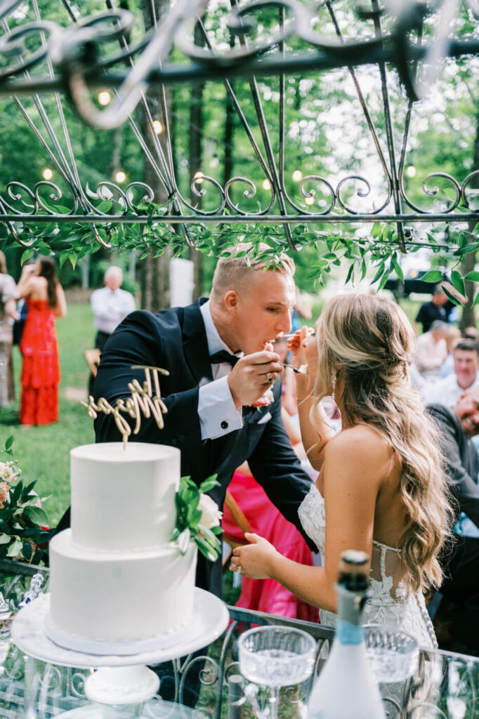 Bride and groom share a romantic cake-cutting moment – The groom in a black tuxedo and the bride in a lace wedding gown feed each other wedding cake under a decorative iron gazebo at their Historic Robertson House wedding.