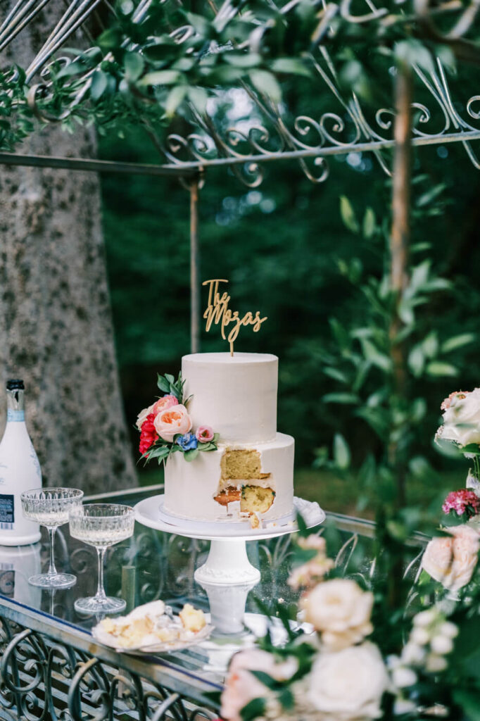 Elegant wedding cake with floral accents – A two-tier white wedding cake with fresh floral accents and a custom topper sits on a glass table at an outdoor wedding reception in North Carolina.