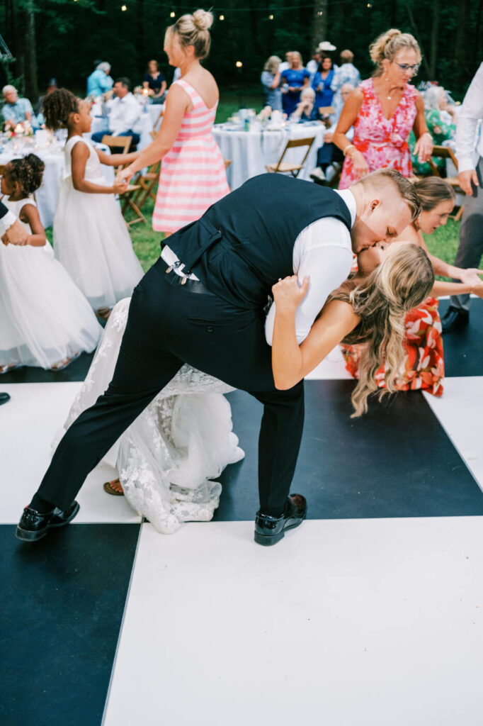 Groom dips bride for a romantic dance floor kiss – The groom, dressed in a vest and tie, leans down for a passionate dip kiss with his bride as wedding guests dance around them.