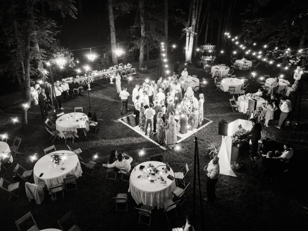 Romantic outdoor wedding reception under chandeliers and string lights – A stunning black-and-white aerial shot captures guests dancing and celebrating at a forest wedding reception in North Carolina.