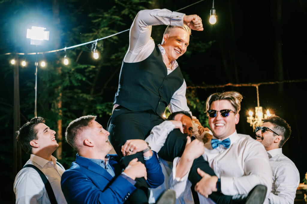 Groom lifted into the air by groomsmen at the wedding reception – The groom is hoisted onto the shoulders of his groomsmen in a moment of celebration, surrounded by glowing chandeliers and string lights.