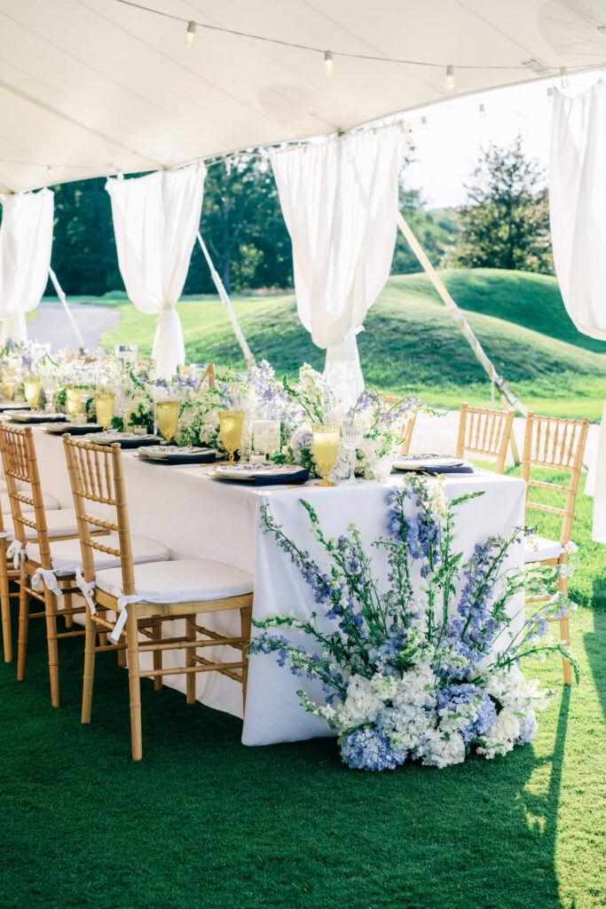 End of a long banquet-style wedding table decorated with loose blue and white florals and golden chairs on green turf